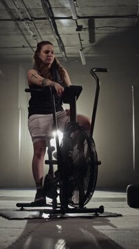 Vertical video of Caucasian young woman resting on air bike after intense workout in gym. Footage is perfect for fitness brand commercials and motivational gym advertisements