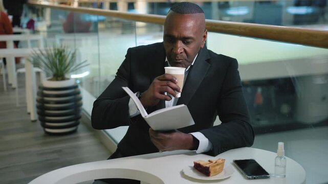 Relaxing African American mature business man drinking coffee with dessert reading book in cafe working pause in company cafe 50s businessman male read dinning with drink at shopping mall work break