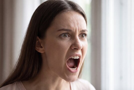 Close-up portrait of a young woman with an angry expression, shouting with wide open mouth and furrowed brow.