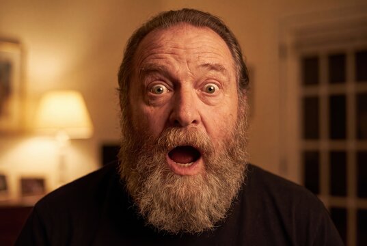 Close-up portrait of a middle-aged man with a thick beard expressing shock and surprise with wide eyes and open mouth.