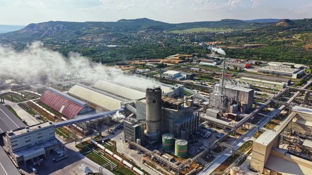 Factory complex features large structures and smoke rising from stacks. Surrounding land is green with hills in the distance. Clear midday sky allows for good visibility.