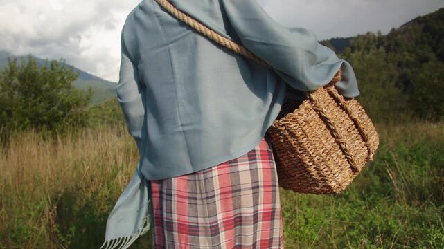 Close-up large wicker basket on shoulder of girl in plaid skirt and blue scarf walking across field. Mountain valley, summer, sunny day. Vacation