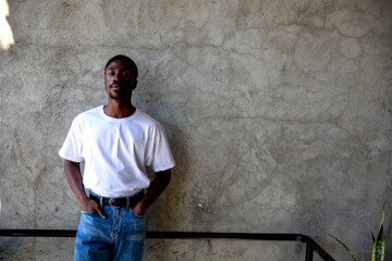 Fototapeta premium Young Black man in a white shirt and jeans standing against a concrete wall