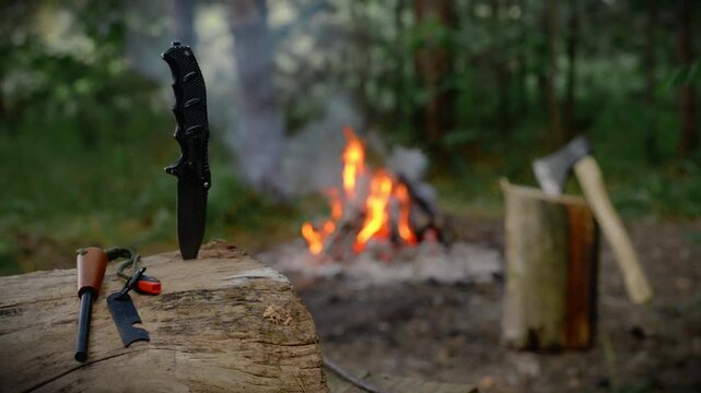 Cinematic B-Roll of Bushcraft Knife Stuck Vertically in Wooden Stump with Burning Fire Background, Crackling Sound, Outdoor Wilderness Survival Gear Video Footage. 