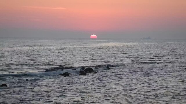 Gry water at sea with ship in horizon, small waves at coast with stones and red sunset and cloudy sky. Topics: natural background, travel, sea ​​view, sea horizon, relaxation, rest, vacation