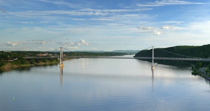 Large suspension bridge spanning across a wide calm Hudson River with forested shores
