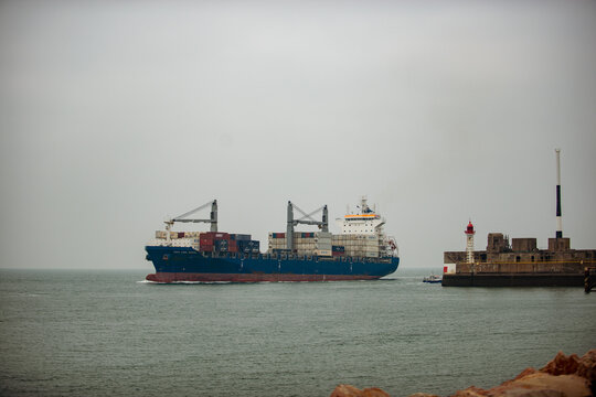 Side view of the container ship "CMA CGM White" heading toward a stone jetty with a red-and-white lighthouse, a tugboat nearby, under an overcast sky at Le Havre.