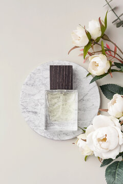 Transparent glass perfume bottle mockup on a round marble podium, decorated with decorative roses and green leaves on a pastel neutral background
