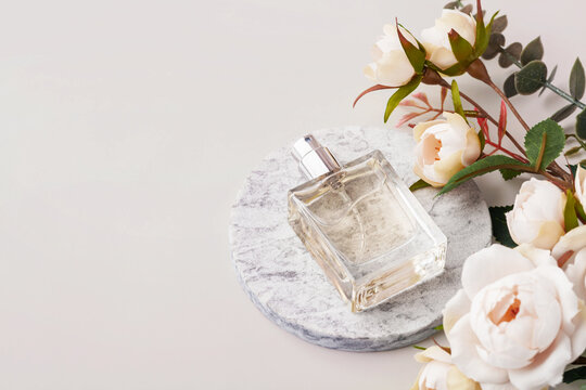 Transparent glass perfume bottle mockup on a round marble podium with floral decorations on a pastel neutral background