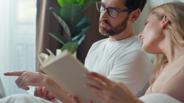 Happy Caucasian married couple relax in bed with computer and read novel. Caucasian man asking help showing laptop discuss online problem with woman reading book. Two partners talk family at morning