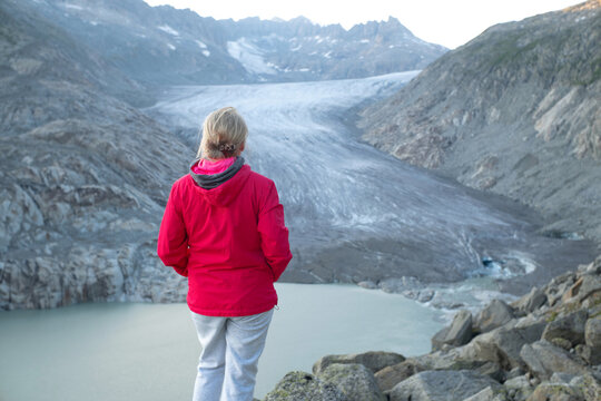 Rear view of an unrecognizable woman in a red jacket standing above a glacial lake and looking toward Rhone Glacier in a rugged alpine valley in Switzerland