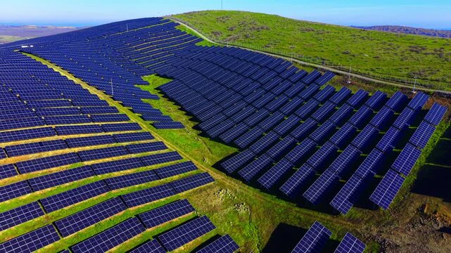 Aerial view of solar array covering green hillside. Rows of dark panels stretch toward a fenced road and open countryside.