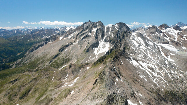 Expansive aerial view of jagged mountain ridges with rocky slopes and lingering snow patches beneath a clear blue summer sky in the Swiss Alps, Switzerland