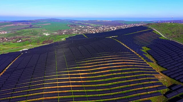 Wide aerial view of solar farm above rural village, Bulgaria. Rolling green hills and rooftops stretch beyond panel rows.