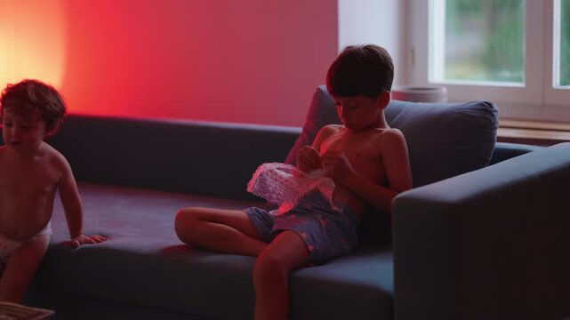 Older brother popping bubble wrap while toddler watches beside him on couch, highlighting sibling bonding, sensory play and quiet family time at home