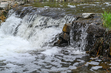 Water cascades over rocky creek edge forming foamy waterfall with spring greenery. Stream creates peaceful outdoor scenery. Waterfall inspires relaxation mood © Olena Vasylieva