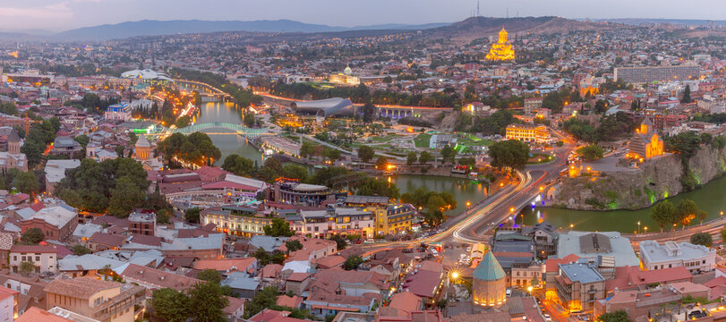 Panoramic view of Tbilisi city center with illuminated bridges, churches, the Kura River and modern architecture at dusk in Georgia.