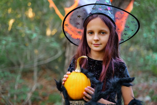 A girl stands in the woods wearing a witch costume. She holds a pumpkin and has a friendly expression. The background has trees and Halloween decorations