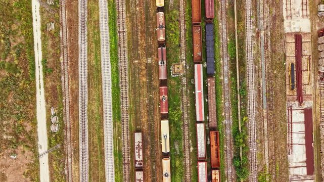 Train cars are parked on tracks in a rail yard. Some cars are rusty and old while others are newer. The scene shows the arrangement of multiple tracks and green areas nearby.