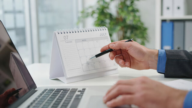 Person pointing at july month on calendar desk planner in modern office.