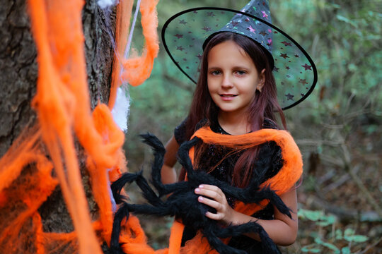 A girl with long hair wearing a witch hat stands next to a tree. She holds a fake spider and is surrounded by orange and black decorations in the woods
