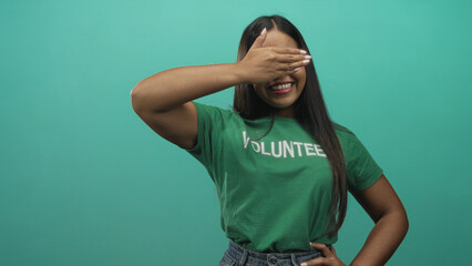 Fototapeta premium Young hispanic woman wearing green volunteer tshirt covering eyes with hand while smiling and hand on hip in studio; community service joy.