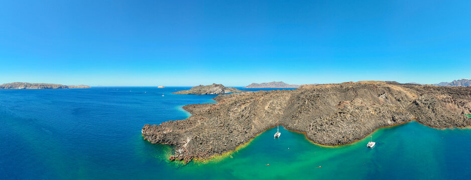 Aerial panorama of a volcanic island surrounded by turquoise waters in the Santorini caldera, Greece, showcasing dramatic geology and coastal scenery.