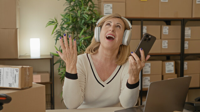 Middle age woman holding smartphone and wearing headphones, hand to chest gesture while talking amid packed boxes in a building; confidence entrepreneurship.