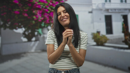 Fototapeta premium Woman clenches hands to chest and laughs with teeth visible on a sunlit street beside white building and pink bougainvillea; joy.