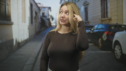 Fototapeta premium Woman pointing finger to temple on street near parked cars and buildings, showing a thinking gesture and puzzled expression; confusion.