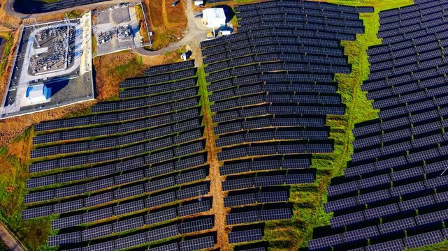 Aerial view of power substation and solar panel. Overhead shot of transformer equipment, service road and panel rows nearby.