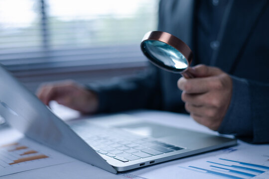 Business professional using a magnifying glass while working on a laptop, representing cybersecurity, digital investigation, online audit, data analysis, business inspection, compliance and risk manag