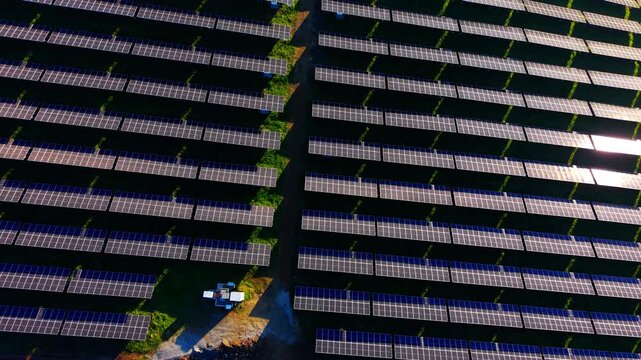 Bright sunlight glinting off solar panel rows. Top down view of parallel arrays with small equipment boxes and dirt path between.