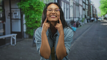 Fototapeta premium Woman points index fingers to cheeks while smiling, wearing yellow top and denim shirt on a street; joy playfulness.