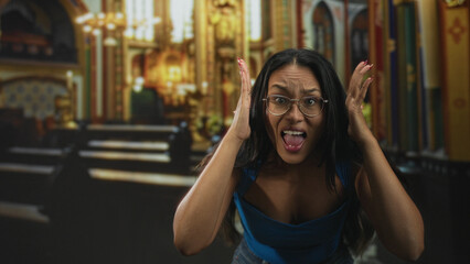Fototapeta premium Young hispanic woman in blue top wearing glasses pressing hands to head and sticking out tongue in ornate church building; surprise playfulness.