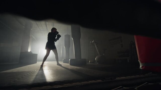 Young female athlete practicing kickboxing, punching and kicking punching bag in dark backlit gym. Clip is perfect for fitness brand commercials and motivational sports advertisements