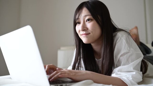 ベッドでノートパソコンを操作して微笑む20代日本人女性
