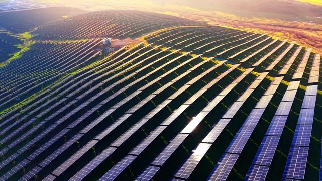 Aerial view of solar farm at sunset. Golden light rakes across curved rows of panels on a hillside with a substation nearby.