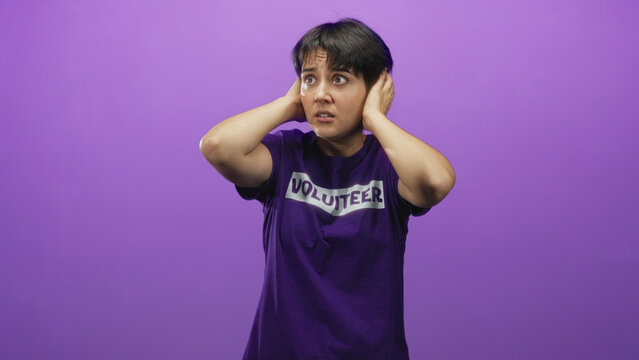 Woman volunteer in purple shirt covers ears with hands and grimaces in a studio while flinching from noise and looking distressed; anxiety overload.