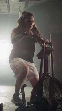 Vertical video of Caucasian young woman training intensely on stationary air bike in gym. Clip is ideal for fitness brands, gym advertisements, and motivational workout campaigns