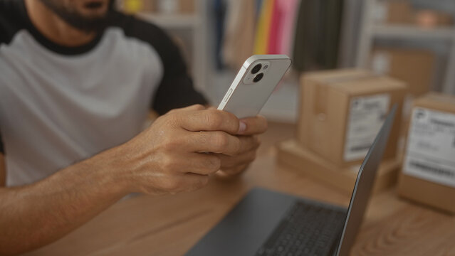 Man tapping smartphone with fingers at packing desk in building, checking orders beside laptop and shipping boxes; small business efficiency focused.