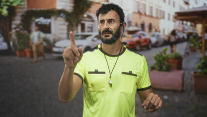 Fototapeta premium Man referee in neon yellow shirt pointing index finger with whistle on necklace and earpiece, beard visible in street; authority control focus.