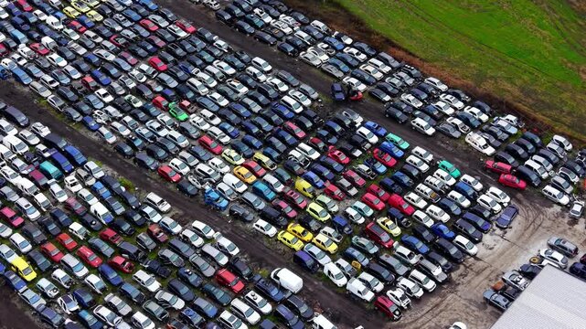 Aerial view of car scrapyard rows. Drone shot of hundreds of parked vehicles in diagonal lines beside green farmland under daylight.