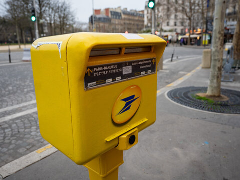 Close-up of a yellow "La Poste" mailbox on a Paris street corner, showing collection details and the postal service's blue arrow logo, with a tree-lined boulevard and traffic light in the background.