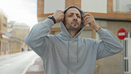 Fototapeta premium Man in gray hoodie with hood up, arms crossed and looking sideways on an empty street beside a building facade; contemplation.