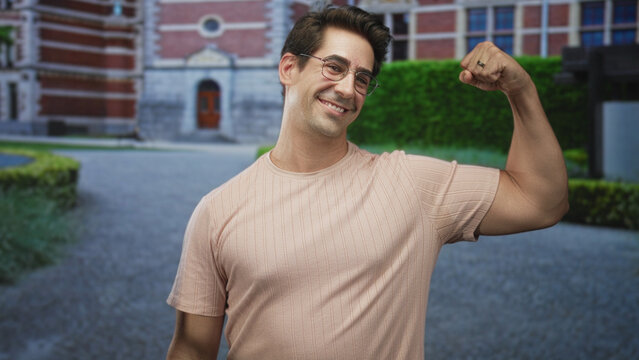 Man flexes bicep and smiles while raising fist in front of a redbrick building entrance, wearing glasses and short sleeve shirt; confidence casual.
