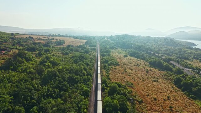 Long freight train travels steadily along centered railway cutting through dense forest and grassy fields toward distant coastal bay under pale blue sky and light haze high elevated viewpoint