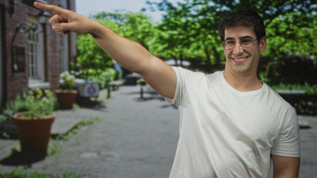 Man in white t shirt and glasses points finger to left with outstretched arm on sunlit street by brick building, large potted plants and leafy park trees; guidance curiosity.