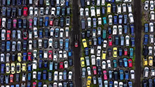 Aerial pattern of scrapped automobiles. Repetitive grid of dented cars in muted colors separated by two straight dirt access roads.
