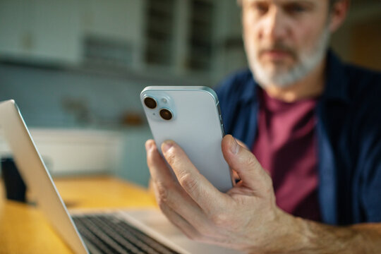 Mature man checking smartphone while working on laptop at home